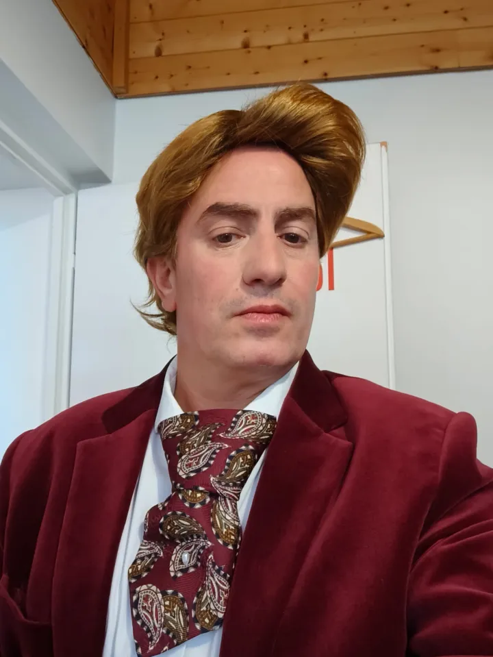 Arthur Green, le présentateur, en veste de velours bordeaux et cravate à motifs, regarde l'objectif de haut